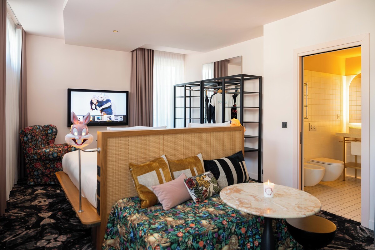Ein Hotelzimmer mit Bett, Fernseher, kleinem Tisch, Stuhl, Kleiderschrank und einem durch eine offene Tür einsehbaren Badezimmer.