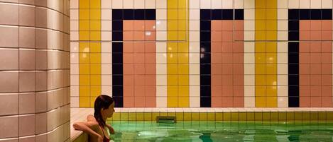 A woman sitting by a pool with tiled walls in a geometric pattern.