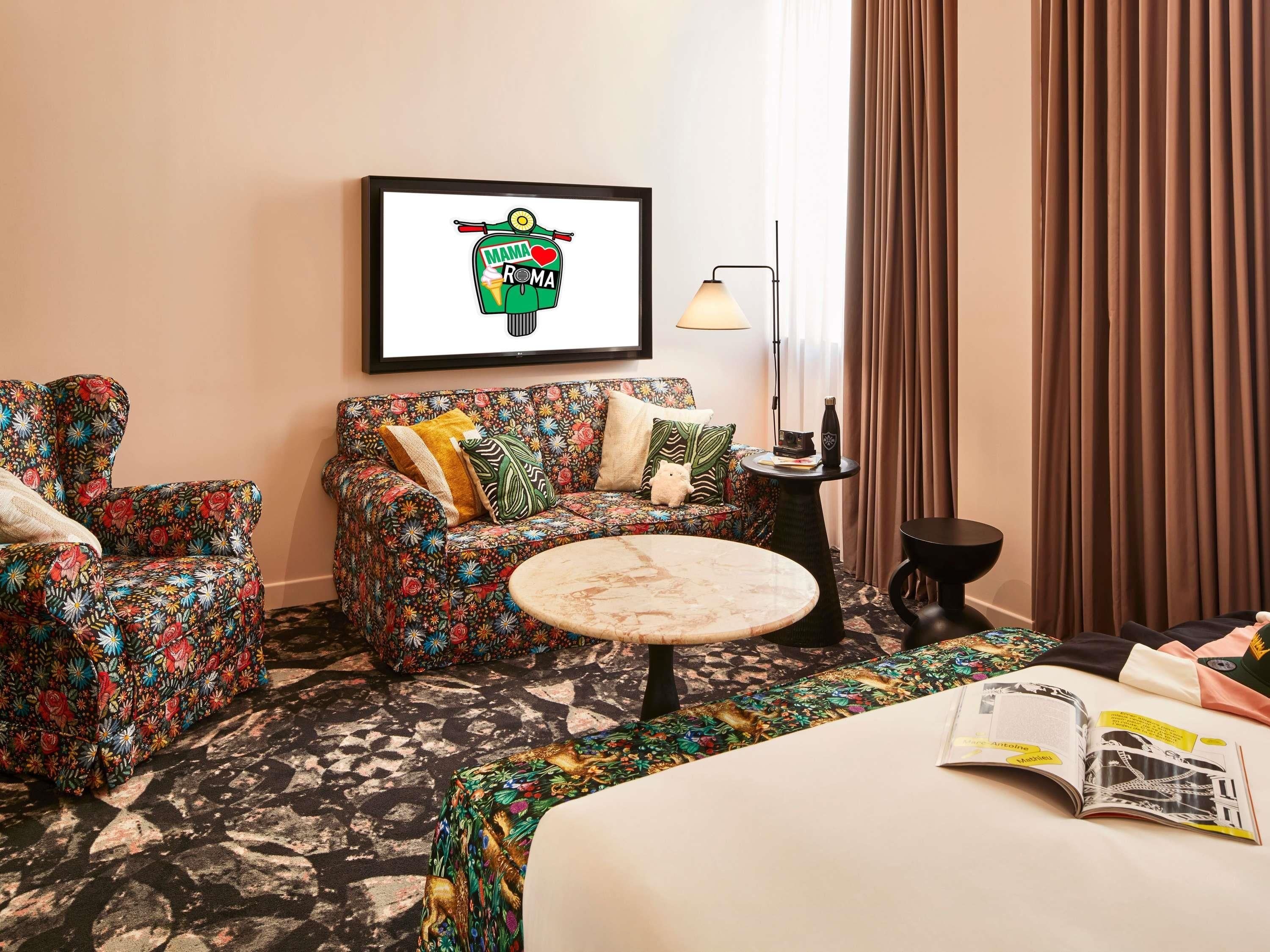 A hotel room with a sofa, armchair, round coffee table, and a bed with a magazine on it.