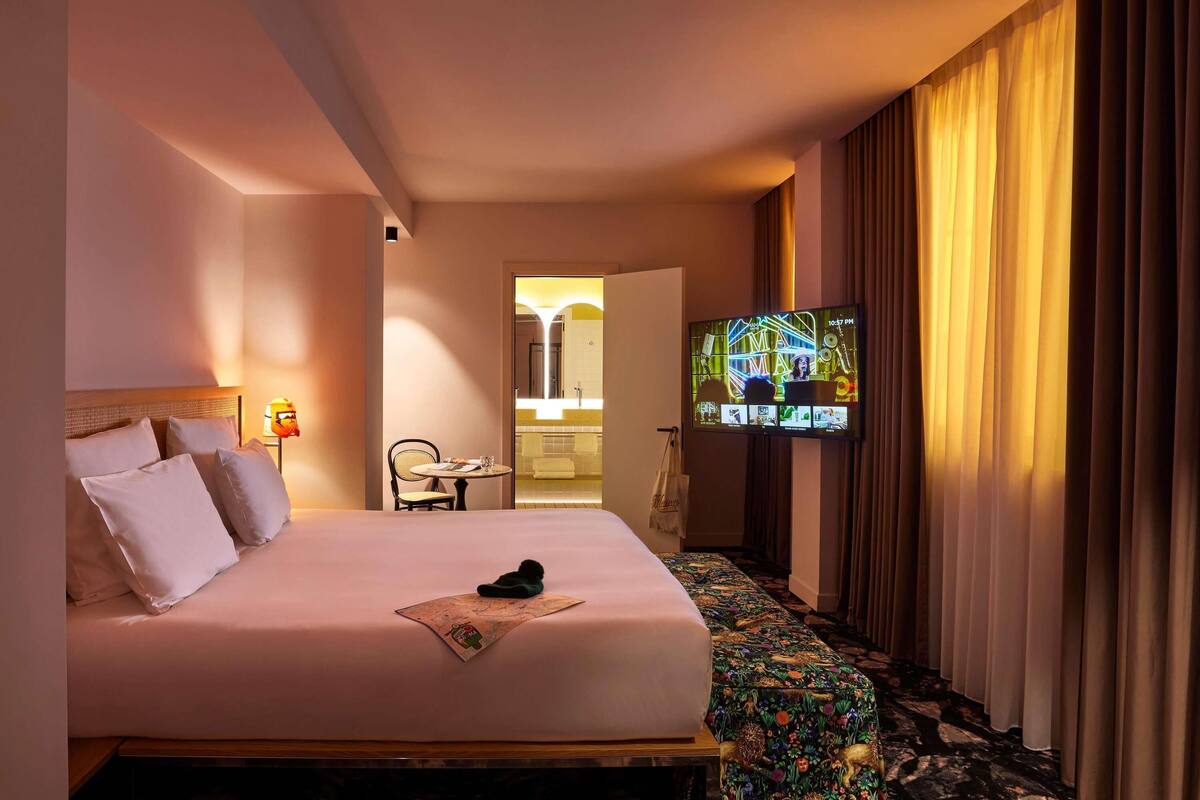 A hotel room with a bed, a television, a small table with a chair, and a view of the bathroom.