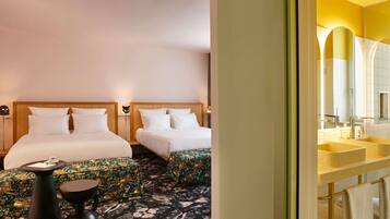 Una camera d'albergo con due letti, un tavolino, un pouf decorato e un bagno con lavandino e specchio.
