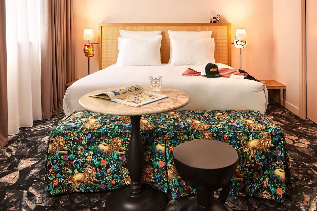 A hotel room with a bed, bedside lamps, a small table with a magazine, a decorative stool, and a patterned ottoman.