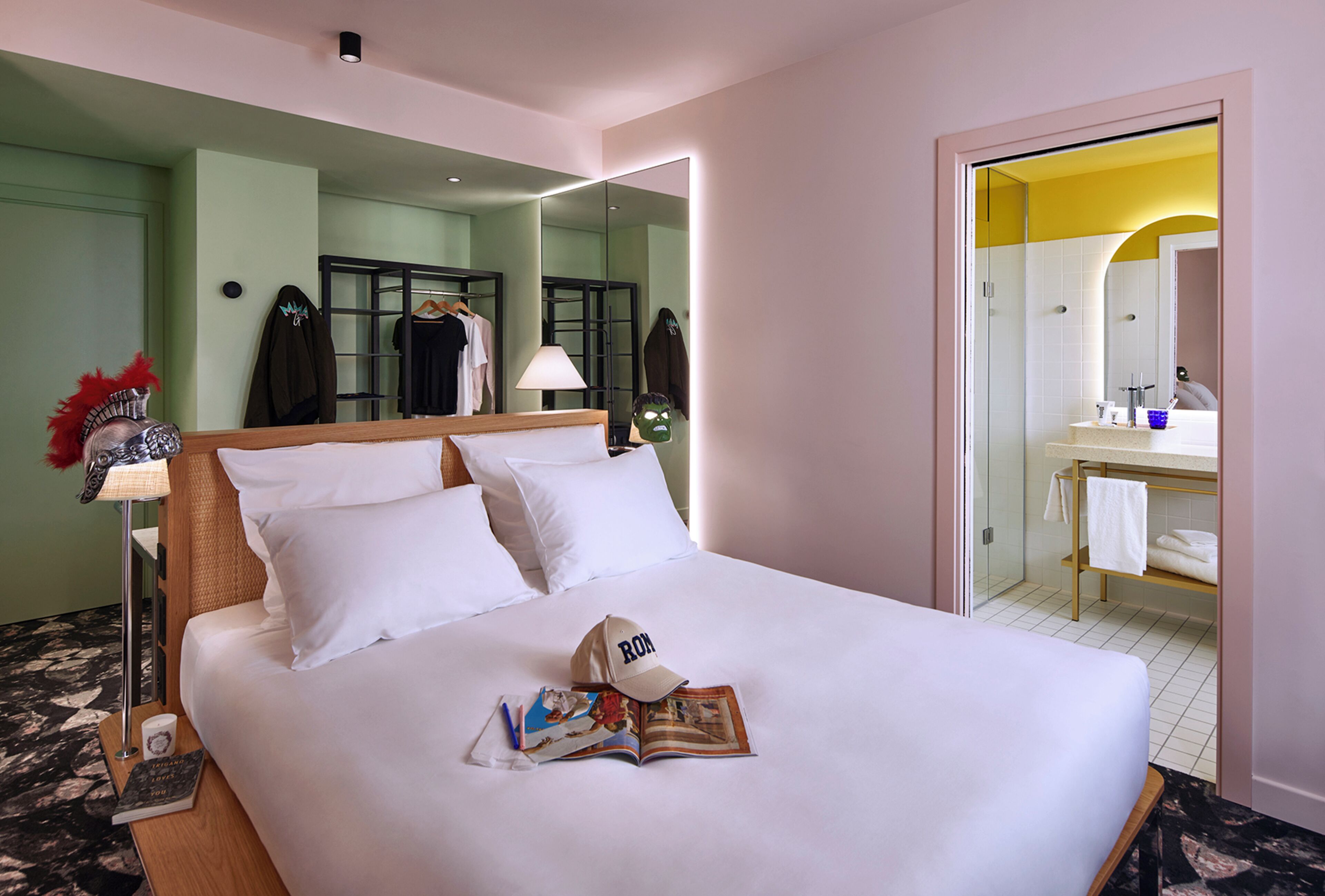 A hotel room with a large bed, a bedside table with a lamp, a mirror, and a view into the bathroom.