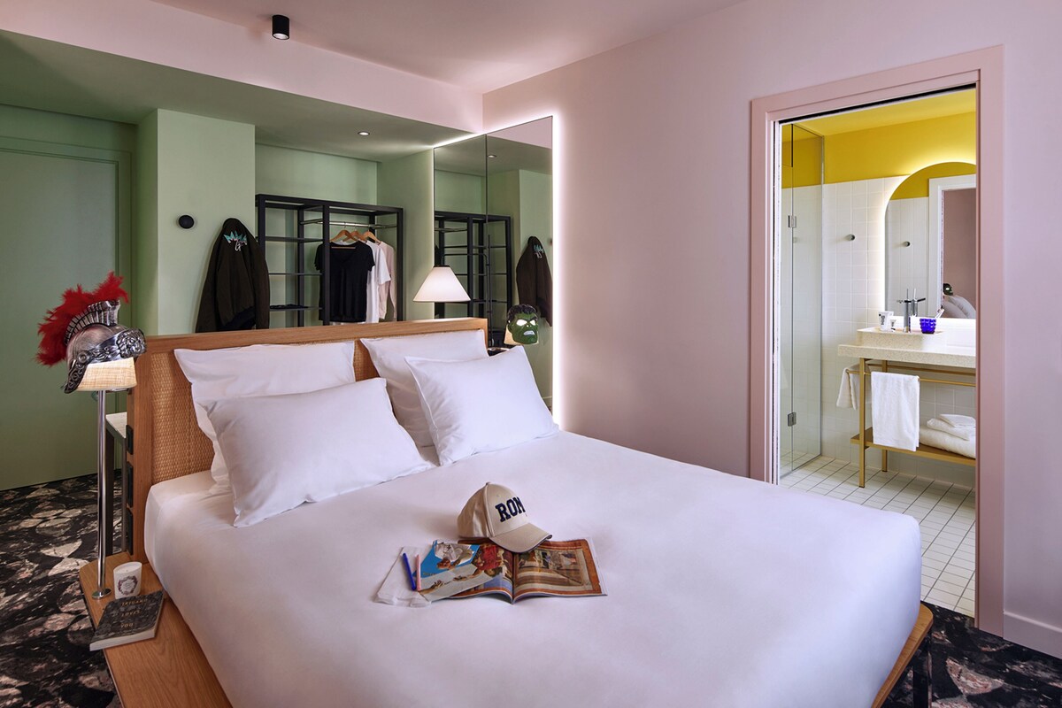 Ein Hotelzimmer mit einem großen Bett, einem Nachttisch mit Lampe, einem Spiegel und Blick ins Badezimmer.
