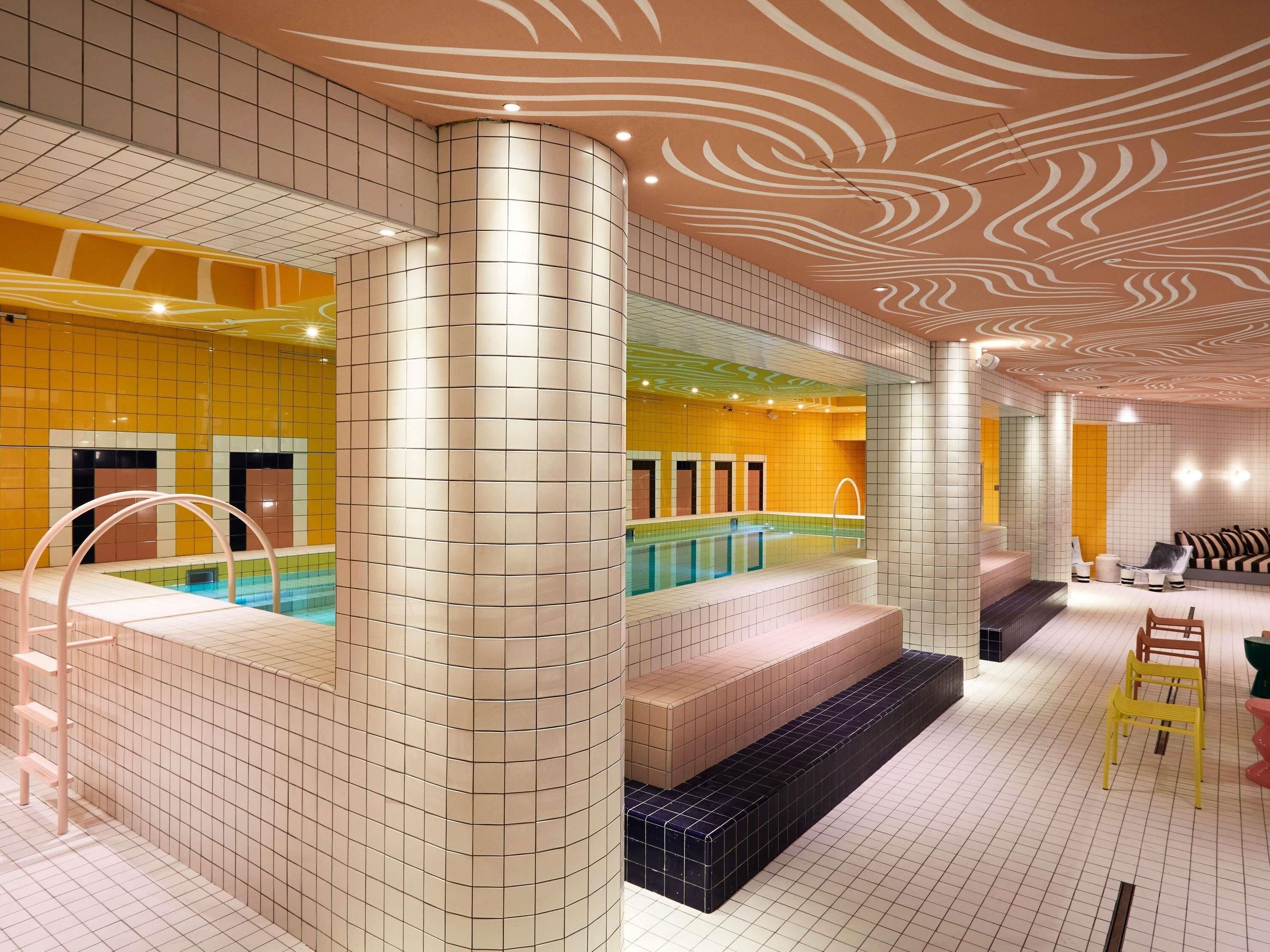 Piscina interior moderna com paredes azulejadas e um pilar curvo, área de estar com sofá e cadeiras, e teto com padrão.