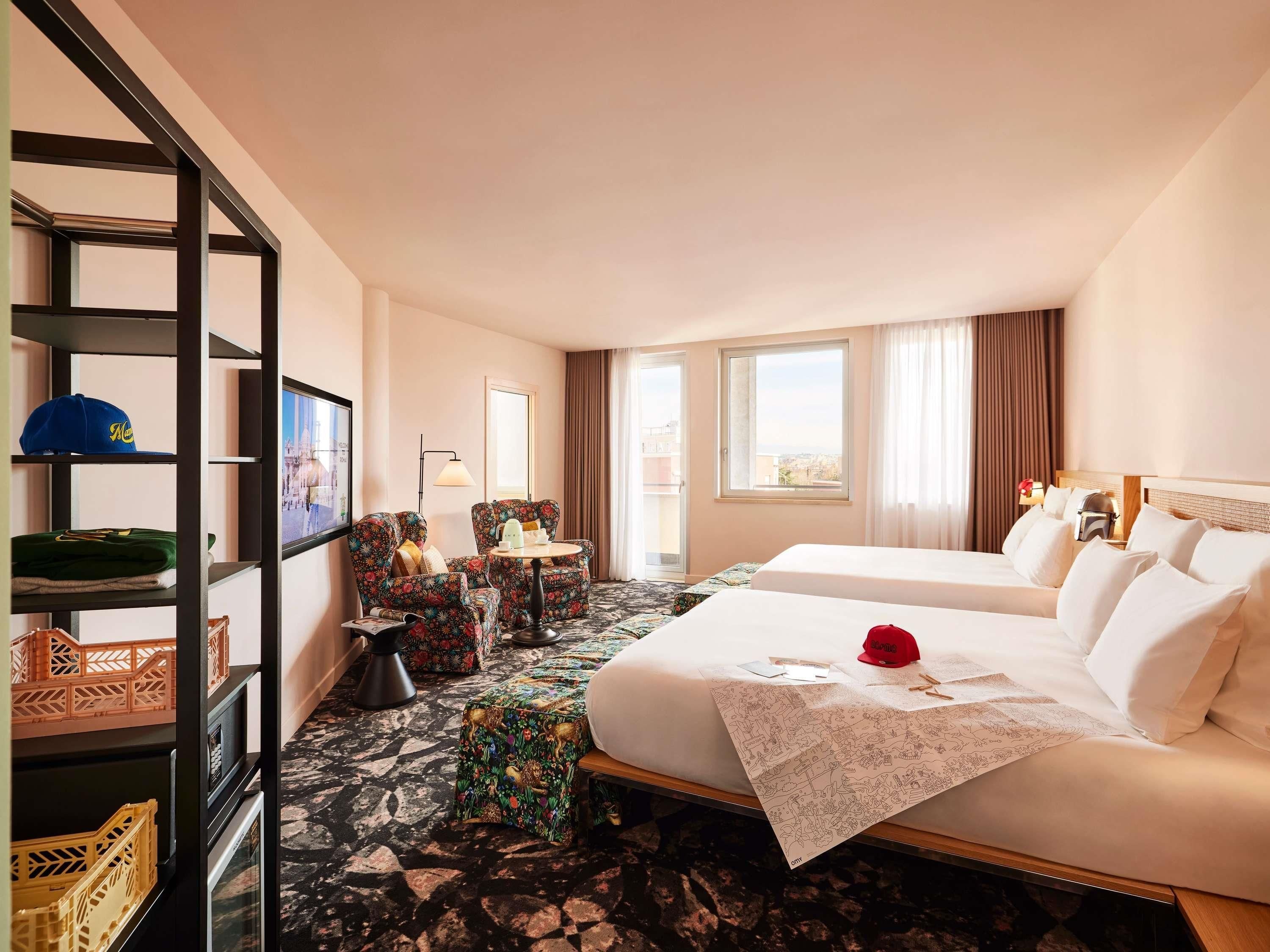 A hotel room with a bed, a desk, a chair, a shelf with hats, and a window.