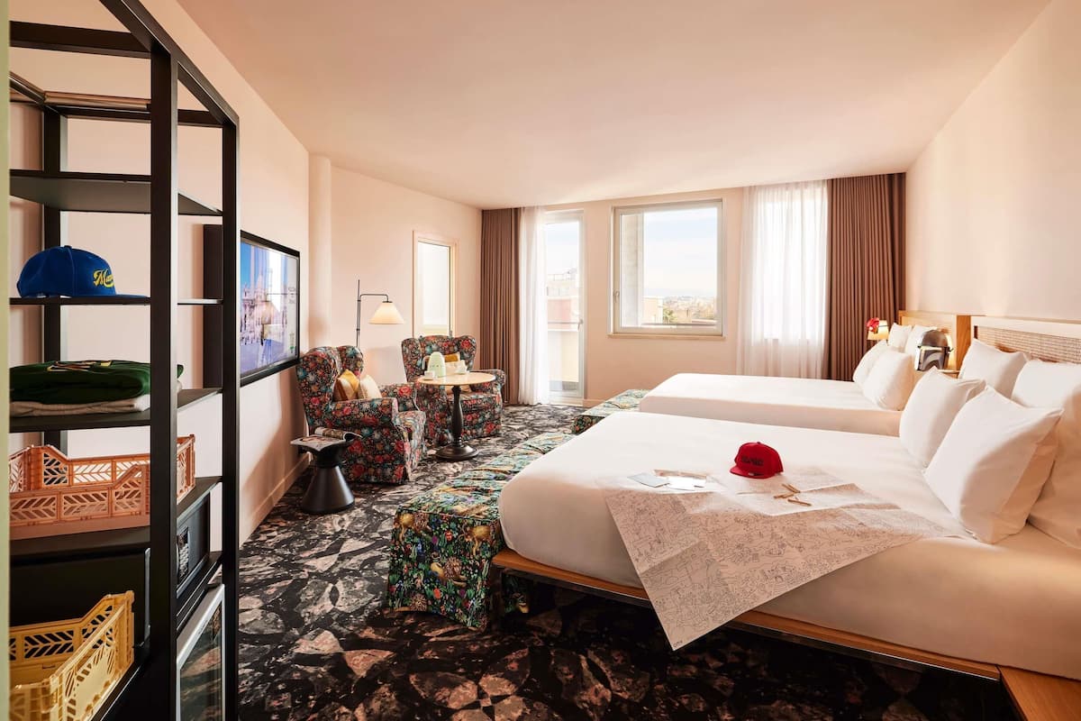 A hotel room with a bed, a desk, a chair, a shelf with hats, and a window.
