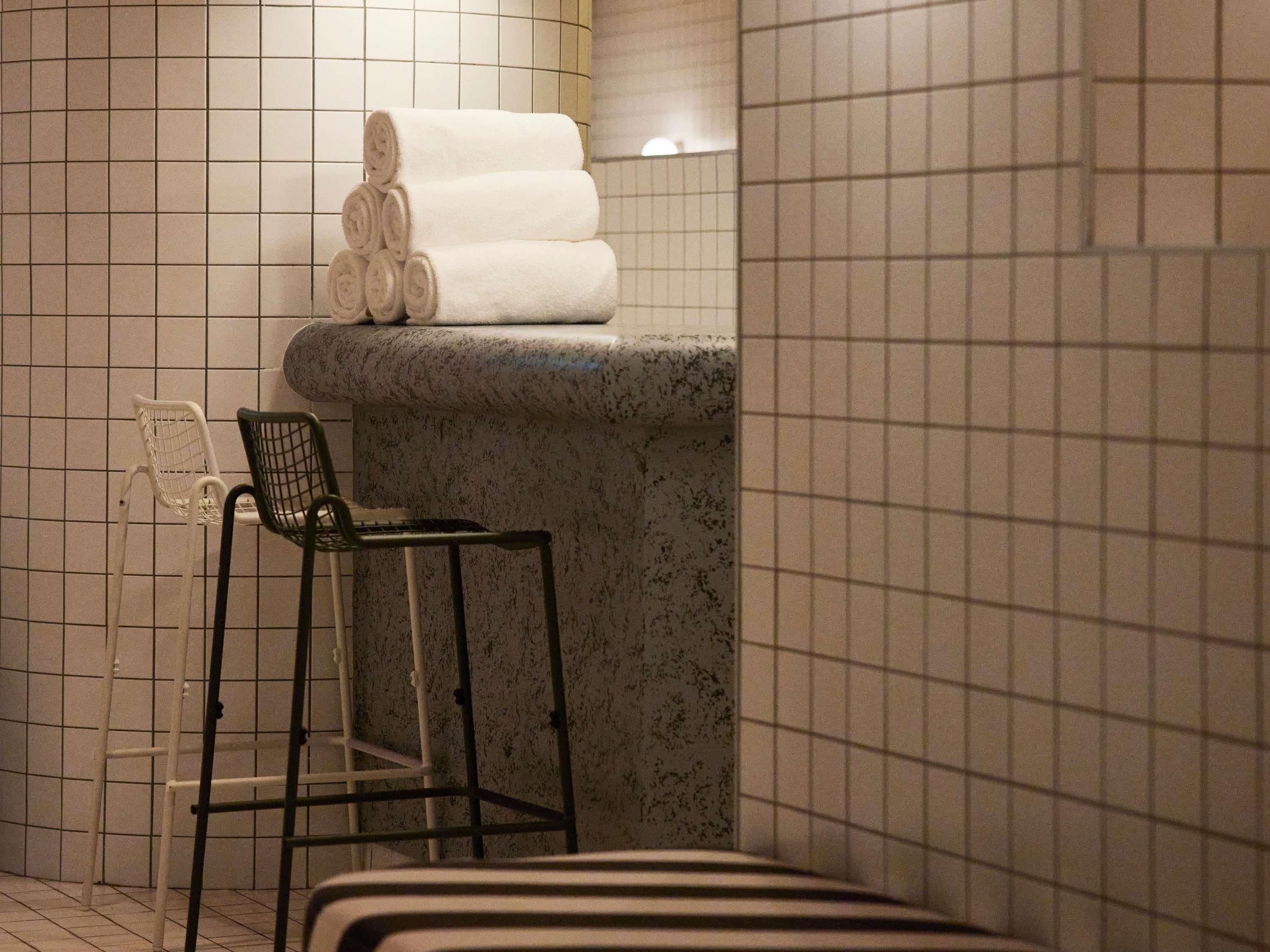 Une salle de bain avec un mur carrelé, un banc en pierre, une chaise et des serviettes enroulées posées sur une étagère.