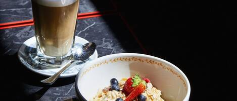 A bowl of cereal with strawberries and blueberries, a glass of coffee, and a spoon on a marble table.