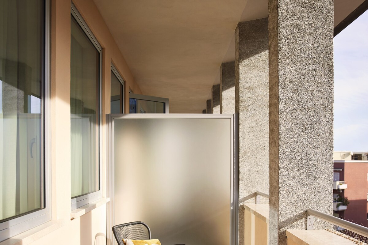 Ein Balkon mit zwei Korbstühlen, einem kleinen runden Tisch mit Früchten und einem Getränk, und Blick auf die Stadtlandschaft.