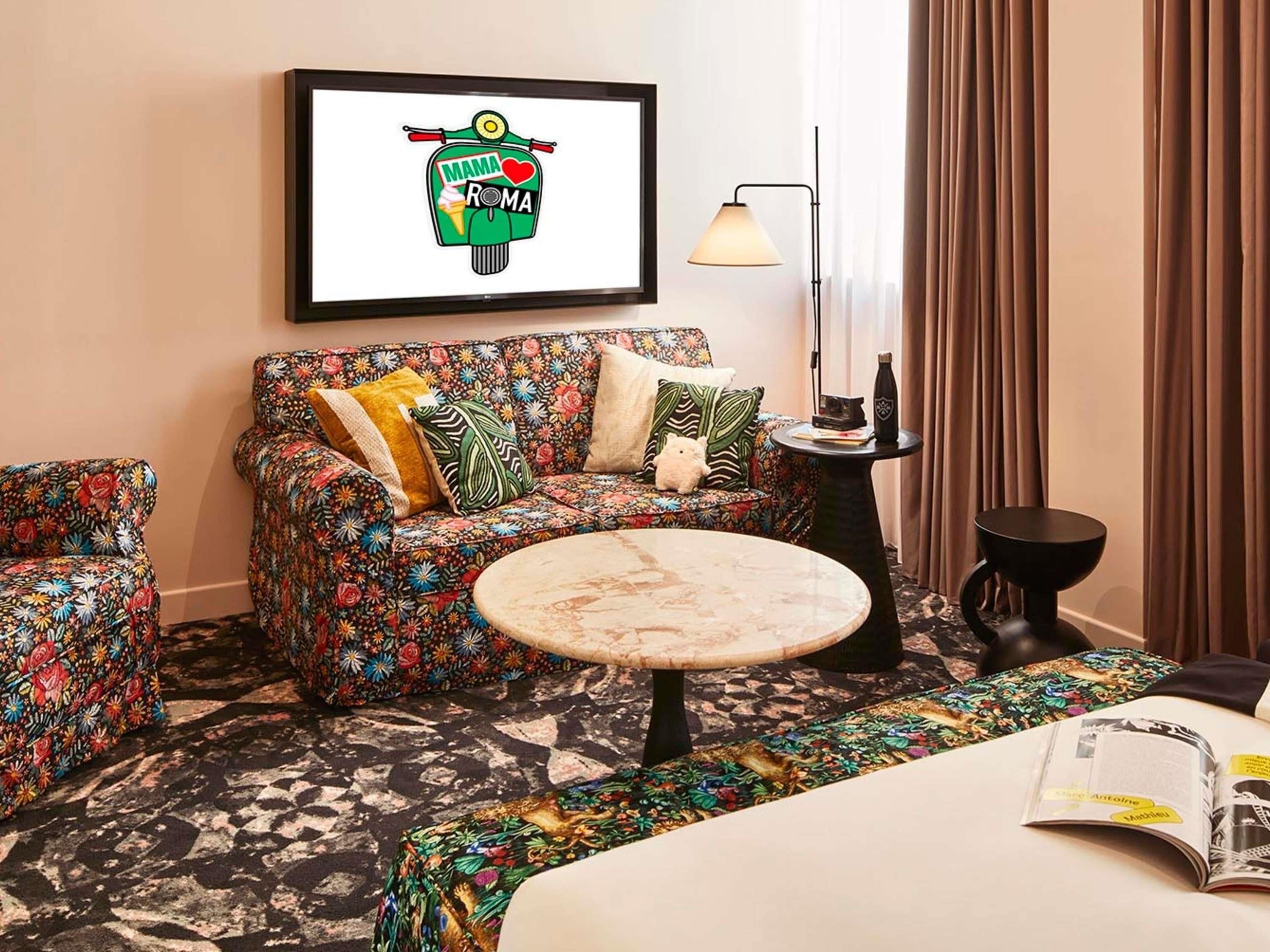 A hotel room with a floral sofa, a round coffee table, a framed picture on the wall, a lamp, and a book on the bed.