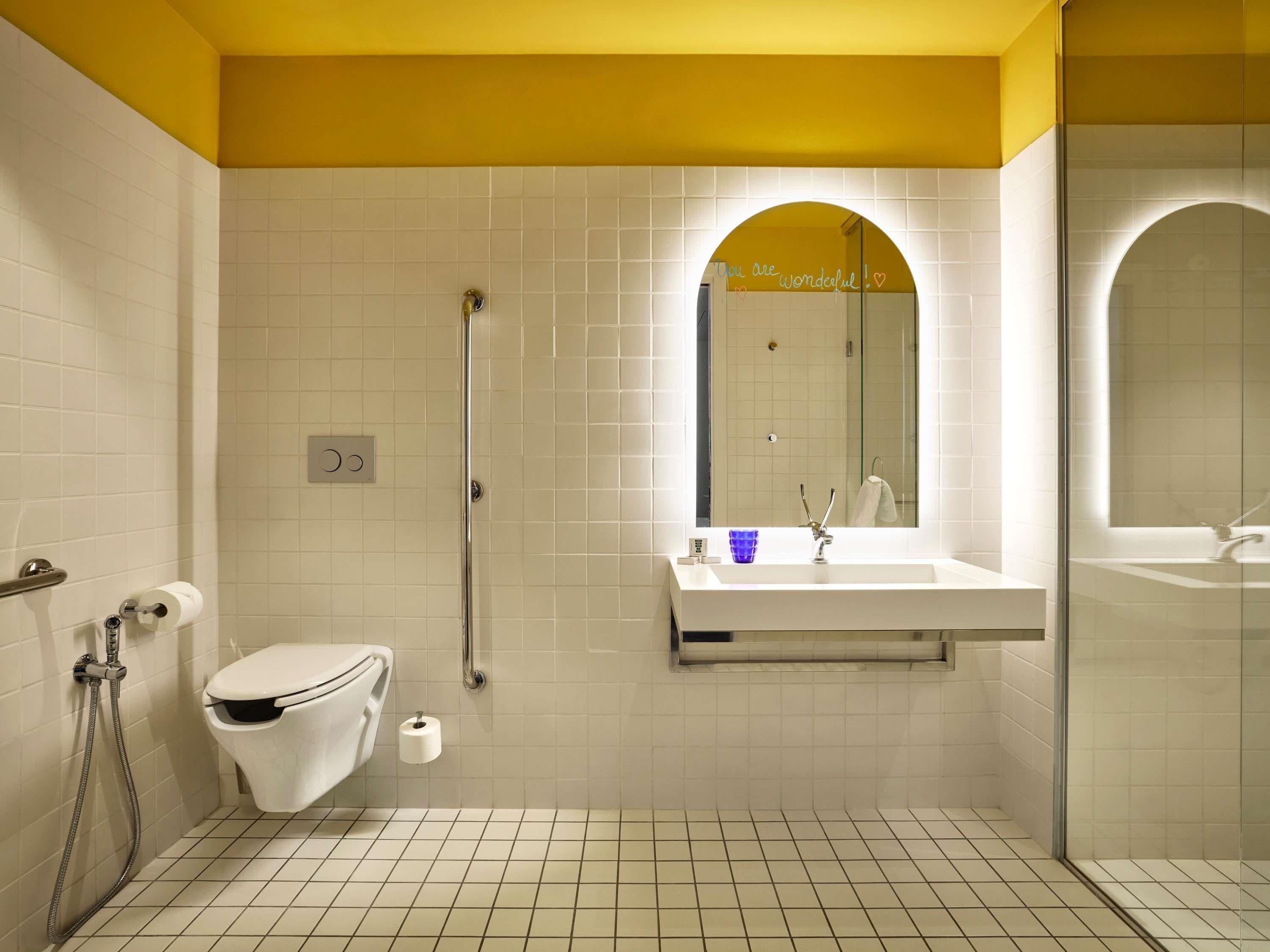 A modern bathroom with a white toilet, a glass shower enclosure, a white sink with a mirror, and a yellow accent wall.