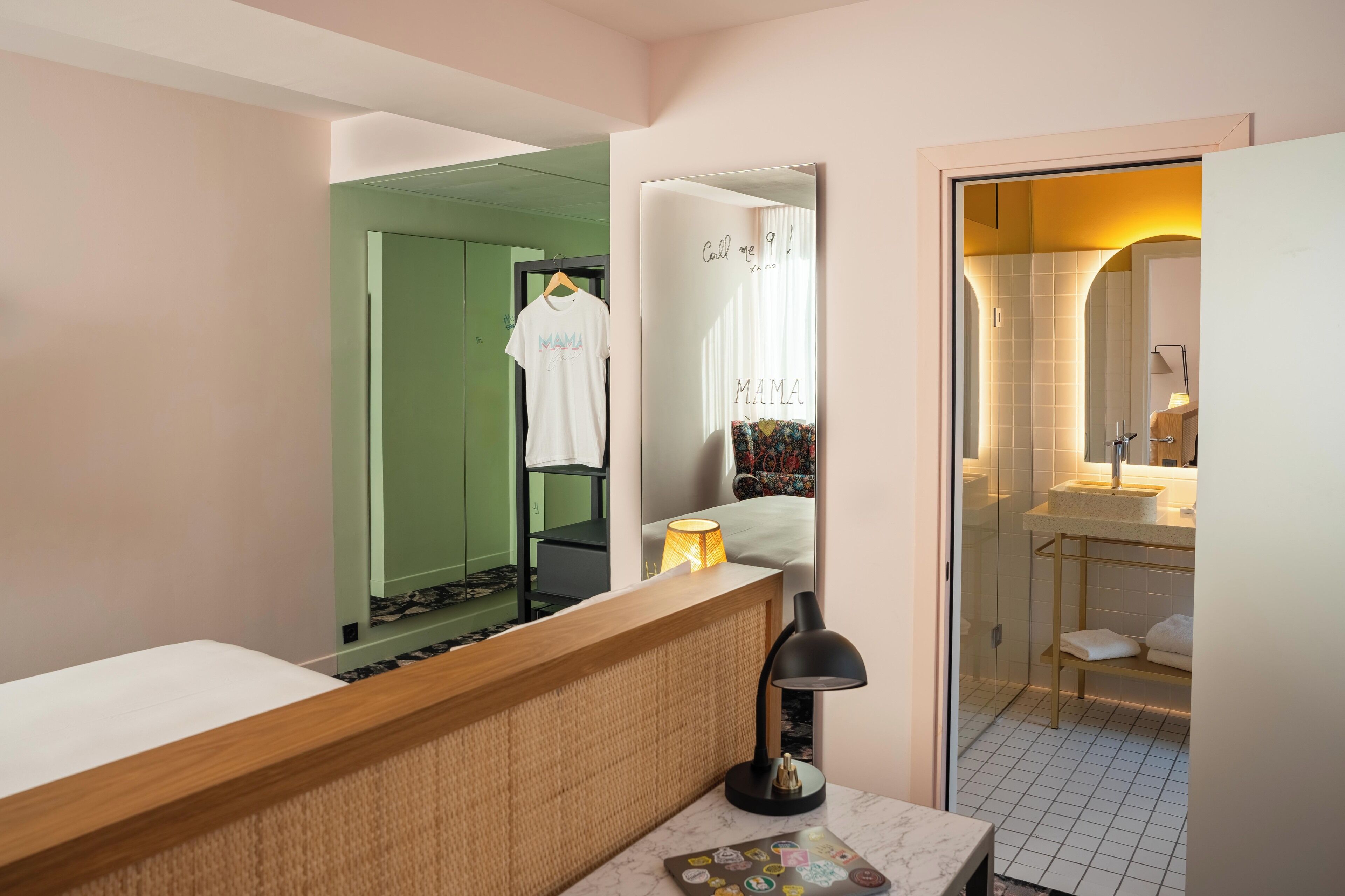 A hotel room with a wooden reception desk, a mirror, a bedside lamp, a white bed, and a bathroom with a sink and mirror.
