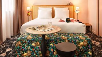 Camera d'albergo con un letto, lampade sui comodini, un tavolino con una rivista, uno sgabello decorativo e un pouf a fantasia.