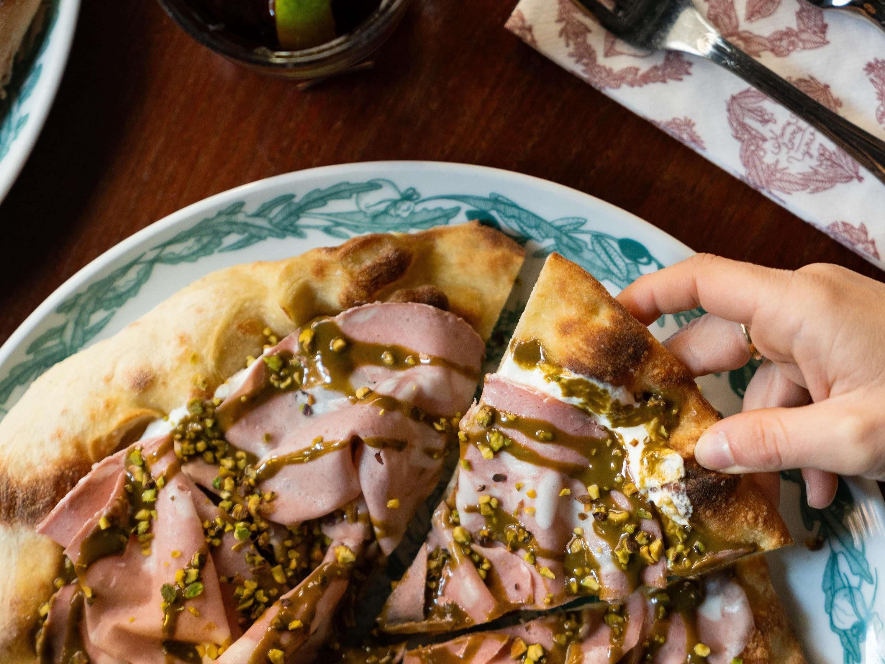 Une assiette contenant une part de pizza garnie de jambon et nappée d’une sauce, posée sur une table recouverte d’une nappe à motifs.
