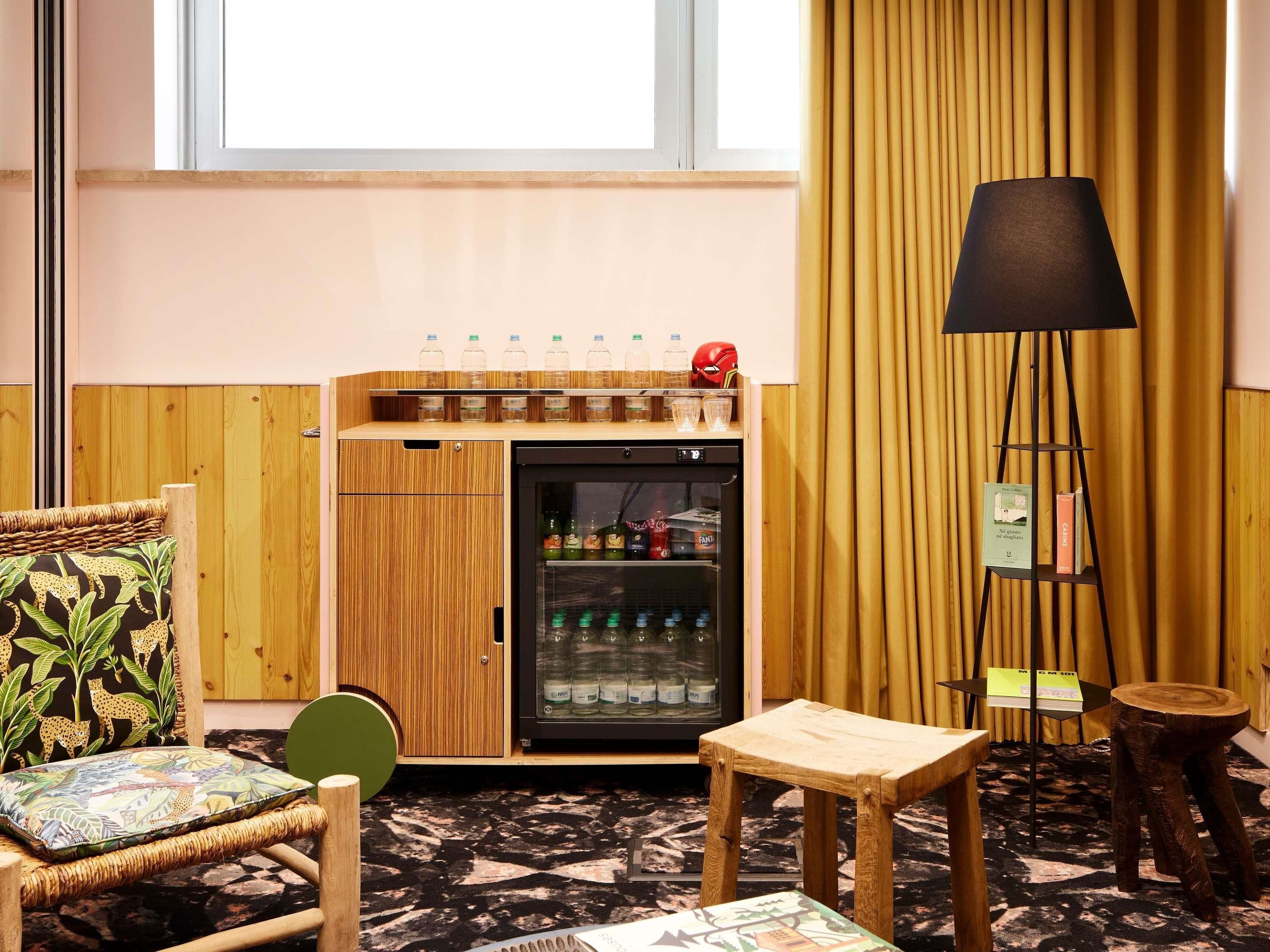 A room with a wooden cabinet, a small refrigerator, a wicker chair, a wooden stool, and a floor lamp.