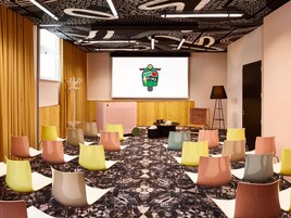 Un salón de conferencias con una pantalla de proyector, varias sillas, un revestimiento de madera y una alfombra estampada.
