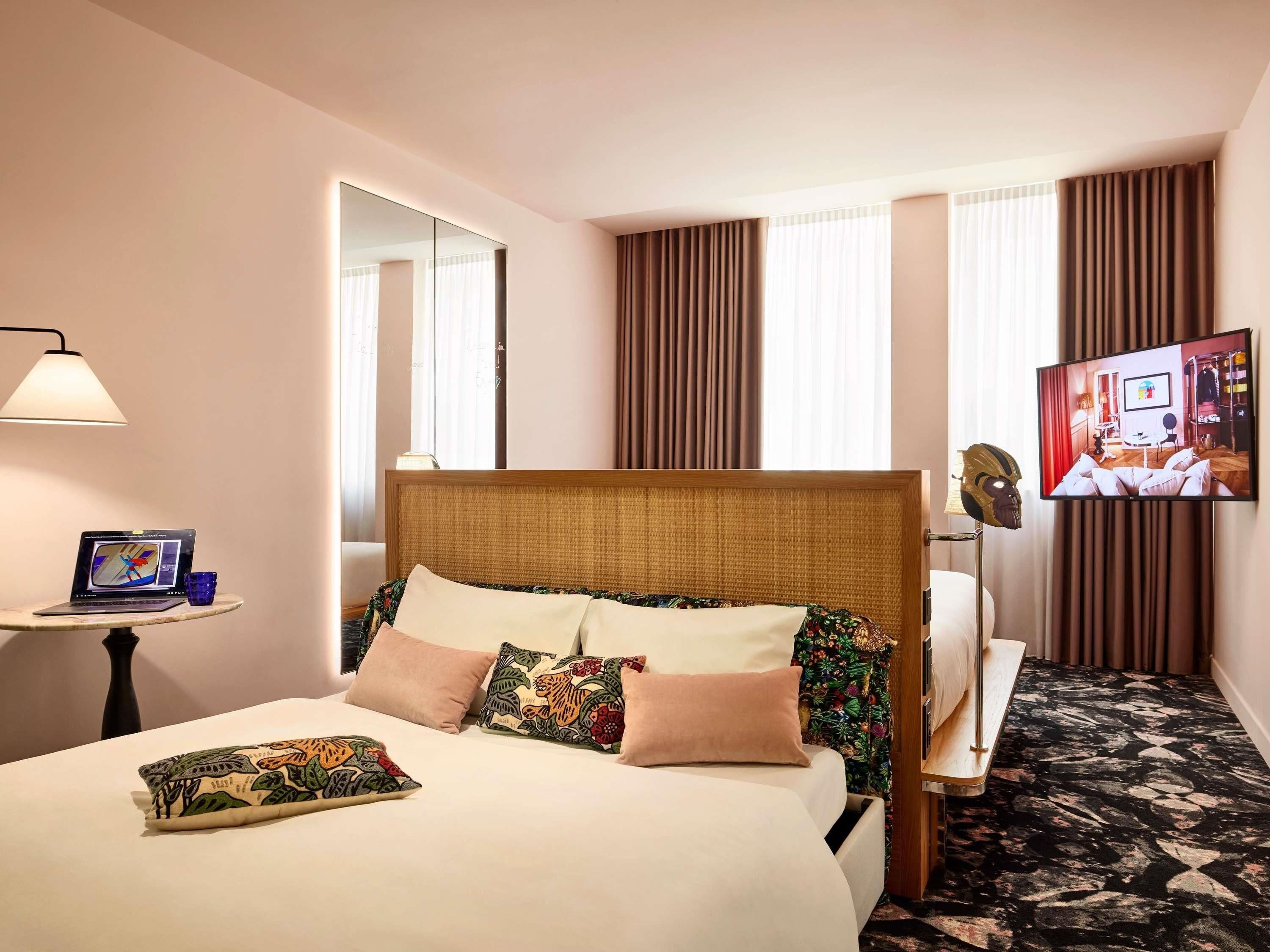 A hotel room with a bed, a TV, a lamp, and a small table with a laptop and glasses.