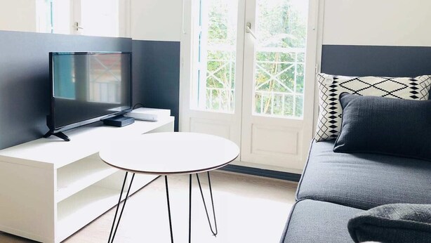 Un salon moderne avec un téléviseur à écran plat posé sur une console blanche, une table ronde blanche et un canapé gris agrémenté de coussins à motifs.