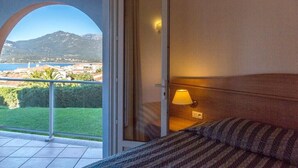 A bedroom with a bed, a nightstand, and a lamp. There is a view of a balcony with a glass door, a mountain, and a body of water in the background. - Residence with sea view (propiano)