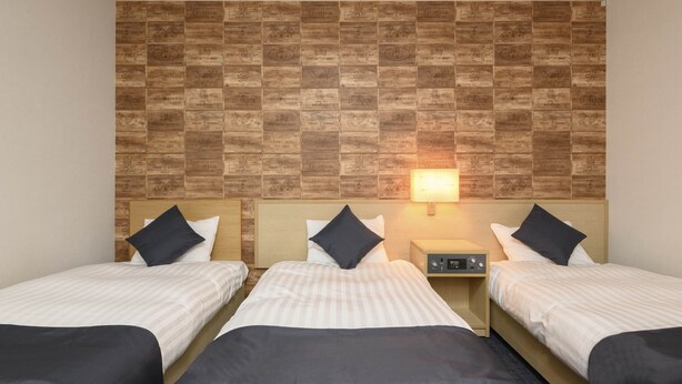 Ein Hotelzimmer mit drei Einzelbetten, jeweils mit einem hölzernen Kopfteil und einem Nachttisch. An der rechten Seite des Raumes ist eine Wandlampe zu sehen.