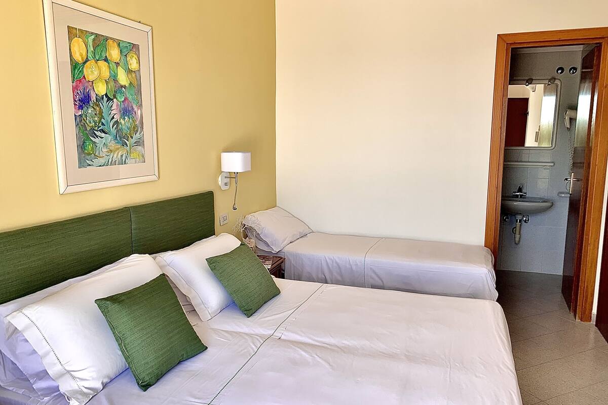 A hotel room with two beds, a painting on the wall, a bedside lamp, and a bathroom visible through an open door.
