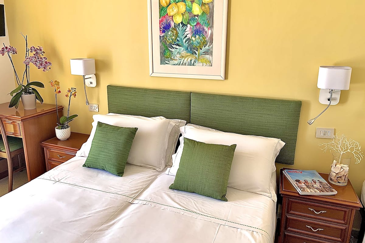 A neatly made bed with green and white bedding, a framed floral artwork above it, and a bedside table with a lamp and a magazine.
