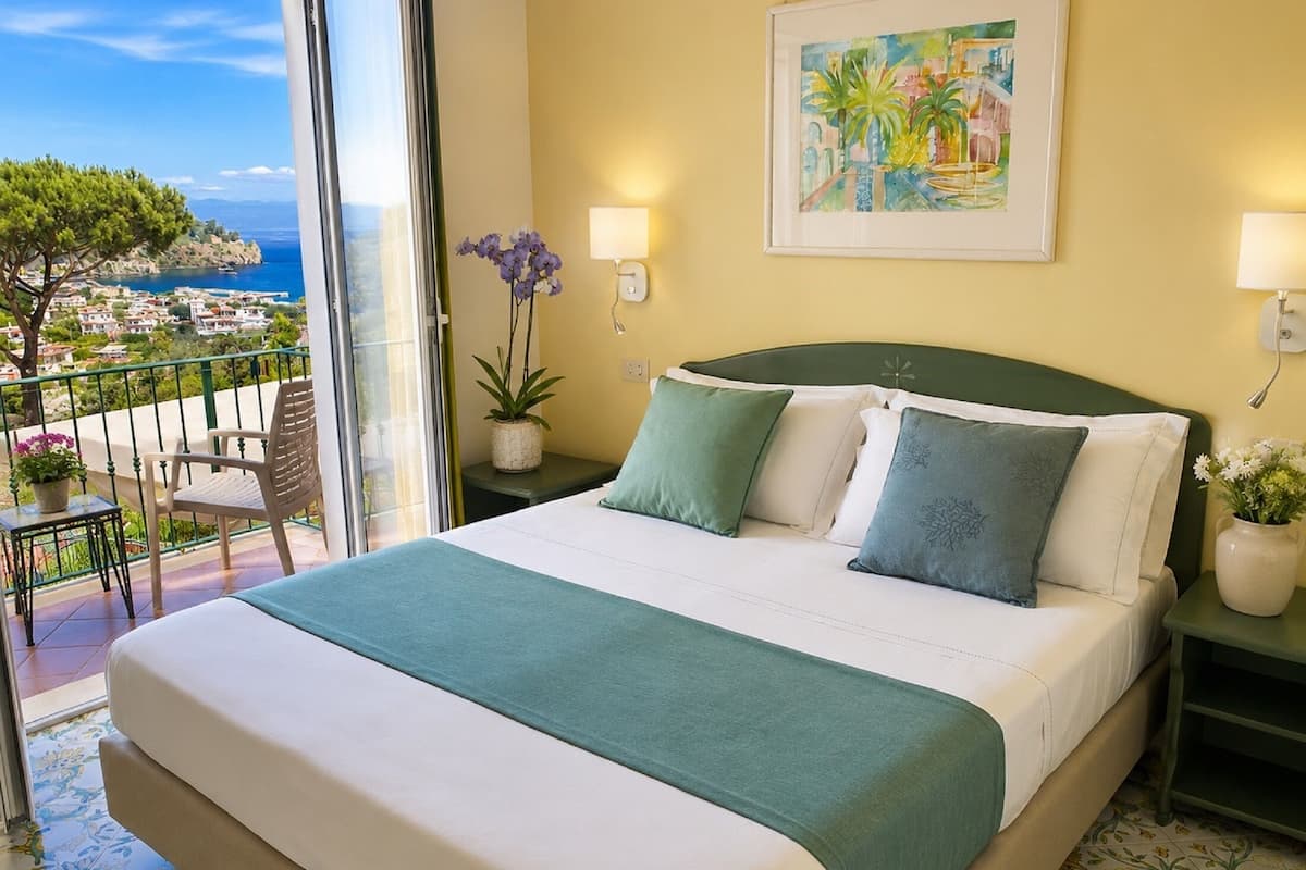 A hotel room with a bed, a view of the sea, a balcony, and a painting on the wall.