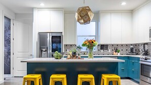 Een moderne keuken met een centraal eiland, gele barkrukken, roestvrijstalen apparatuur en decoratieve planten.