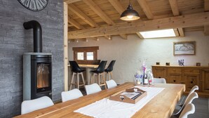Une table à manger en bois, des chaises, une cheminée en pierre et une horloge au mur.
