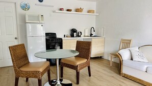 Ett kompakt kök med kylskåp, spis och mikrovågsugn. Det finns en matplats med ett glasbord och flätade stolar. En vit soffa och en stol står längs väggen.