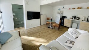 Ett modernt vardagsrum med en platt-TV, en matplats med ett glasbord och ett kök med mikrovågsugn och kaffebryggare.
