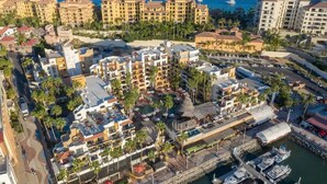 Vista aérea de una zona hotelera costera con varios edificios, un puerto deportivo con barcos y una plaza central con palmeras.