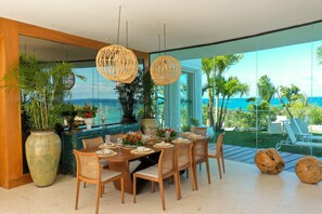 A dining area with a view of the ocean, featuring a wooden table set for six, wicker chairs, and hanging light fixtures.