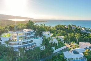 A modern multi-story building with a rooftop pool, surrounded by lush greenery and overlooking a coastal area.
