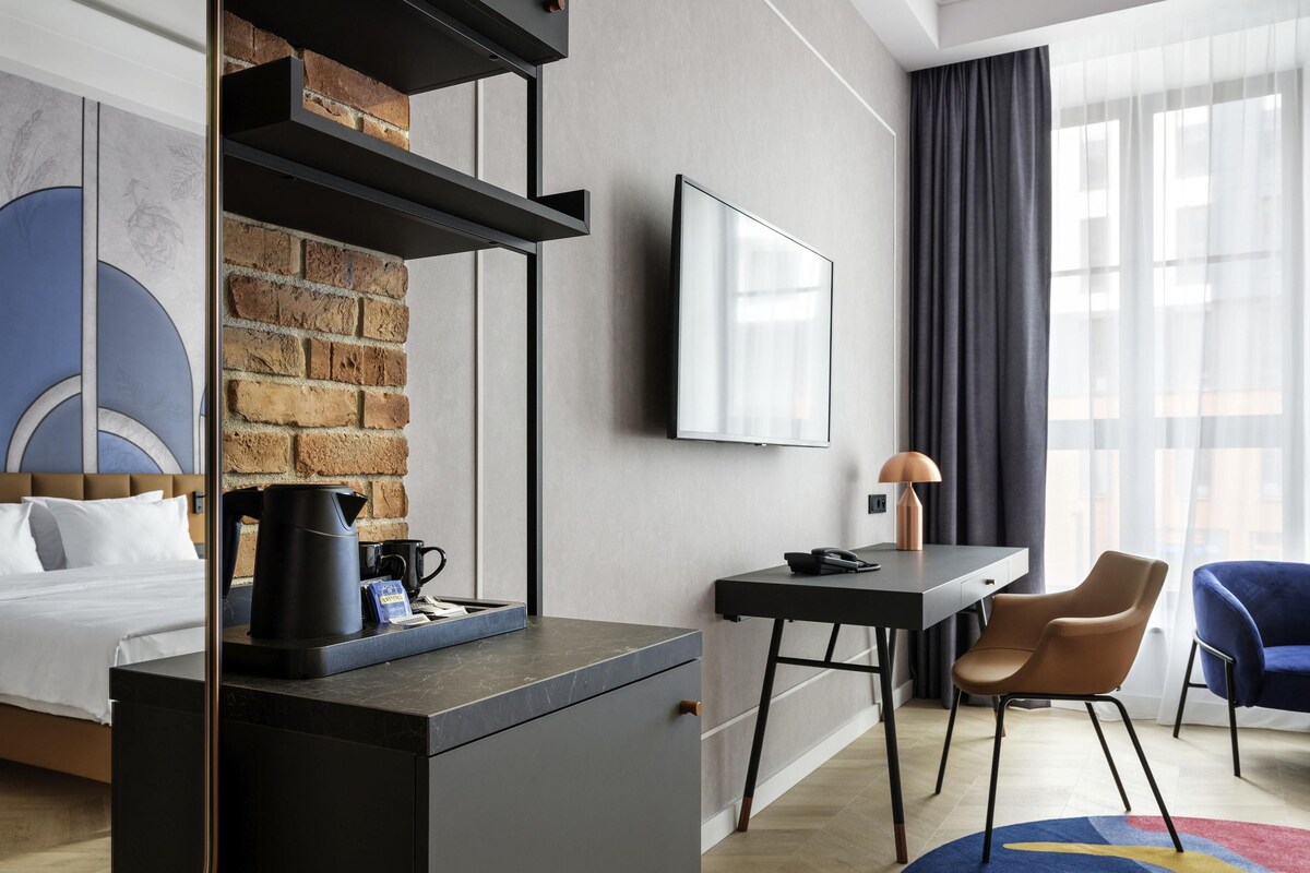 Une chambre d’hôtel moderne équipée d’un lit, d’un bureau, d’une machine à café, d’une télévision et offrant une vue sur la ville grâce à de grandes fenêtres.