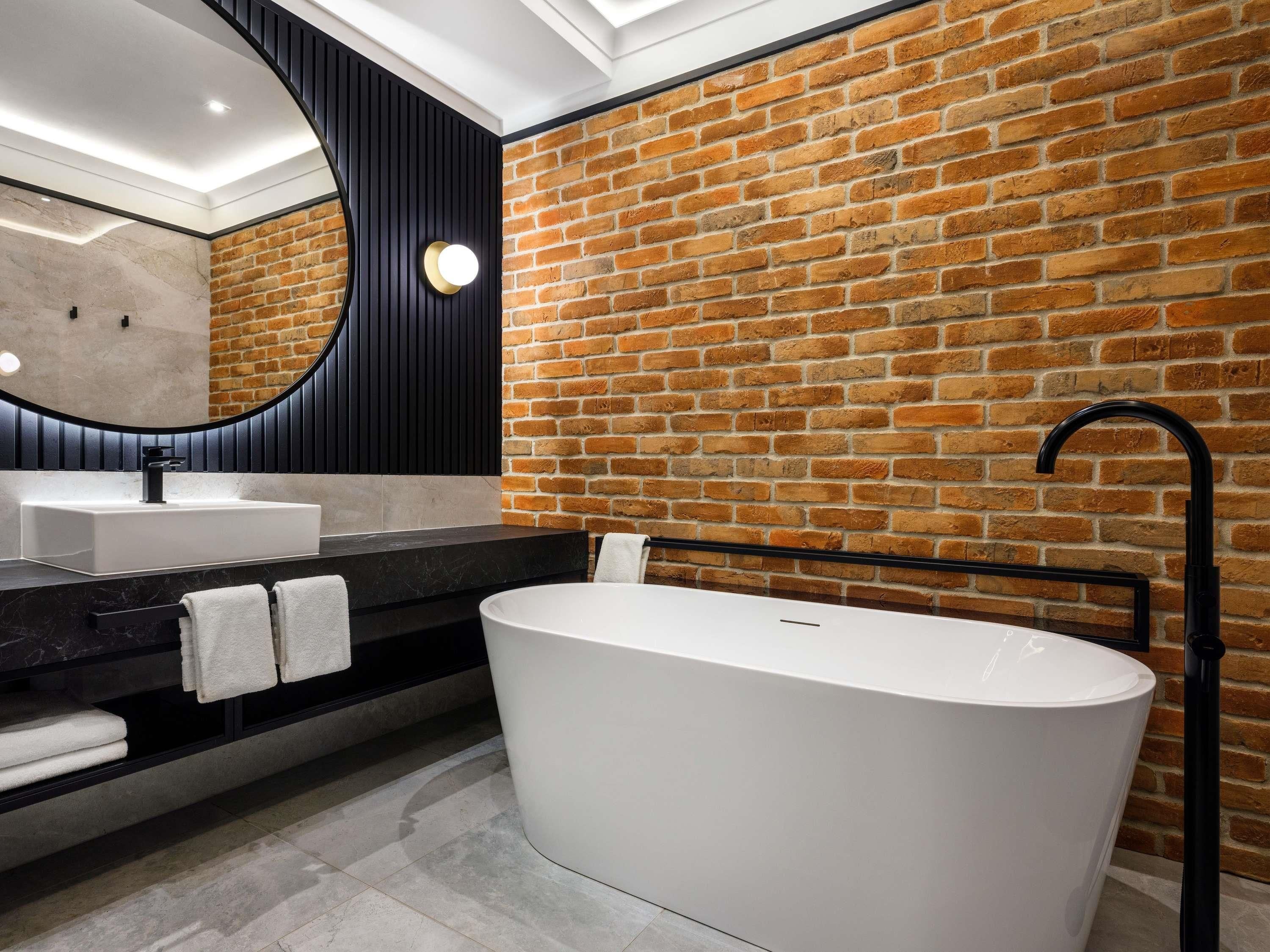 A modern bathroom with a brick wall, a freestanding bathtub, a large mirror, and a black countertop.