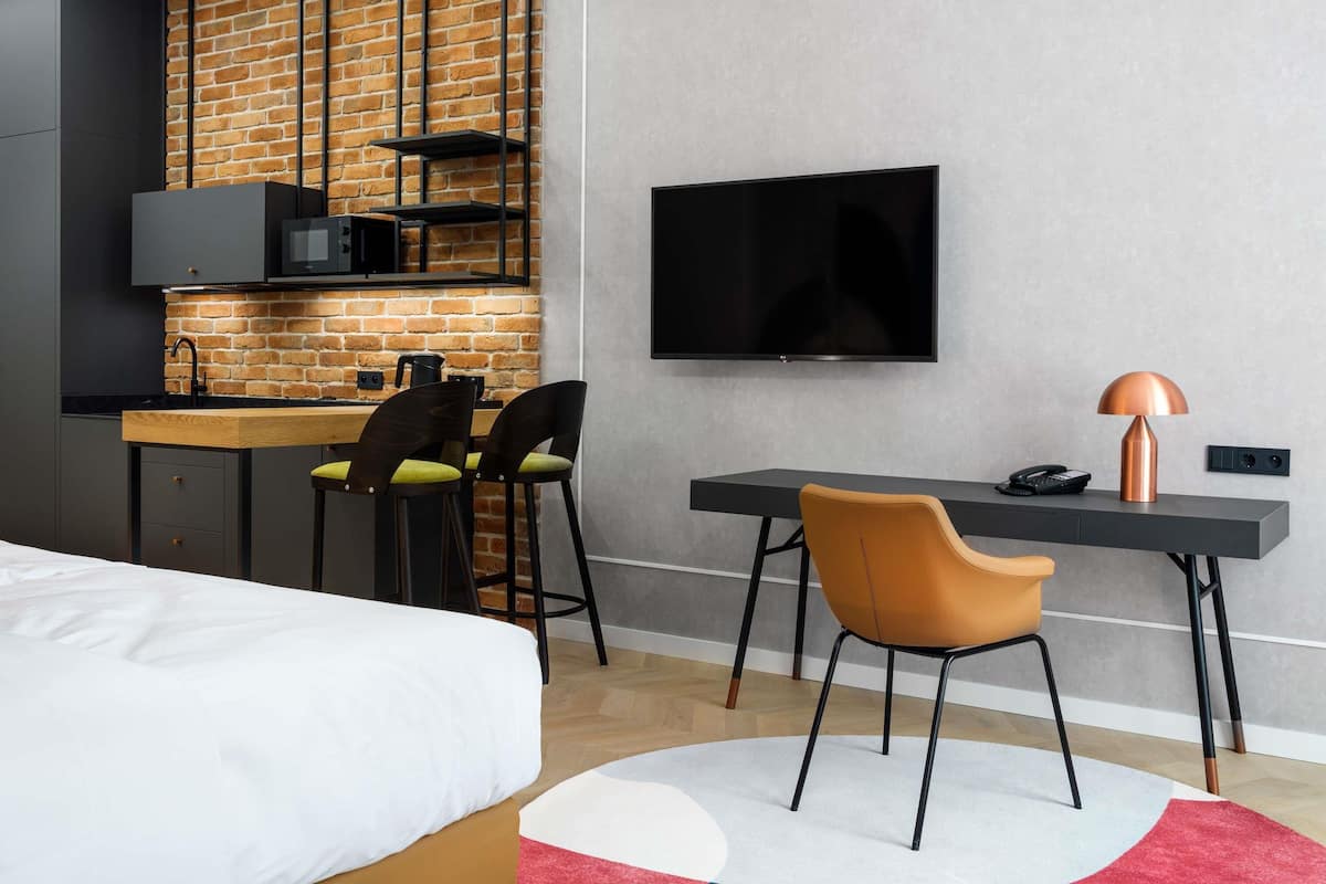 Ein modernes Hotelzimmer mit KĂŒchenzeile, einem Bett, einem Schreibtisch mit Stuhl und einem Fernseher.