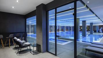 A modern indoor pool area with glass walls, a seating area with chairs and tables, and a ceiling with linear lighting.