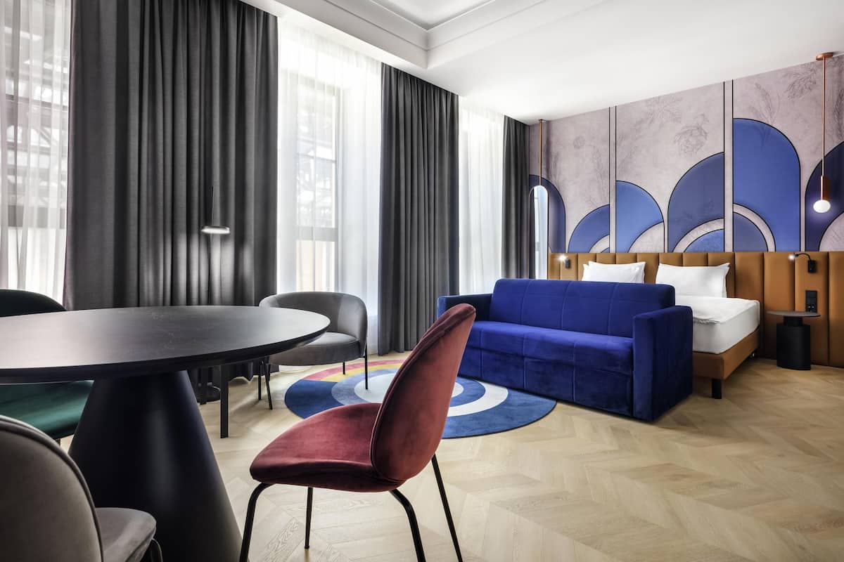 Una habitación de hotel moderna con un sofá azul, una mesa de comedor redonda y un ventanal con cortinas.