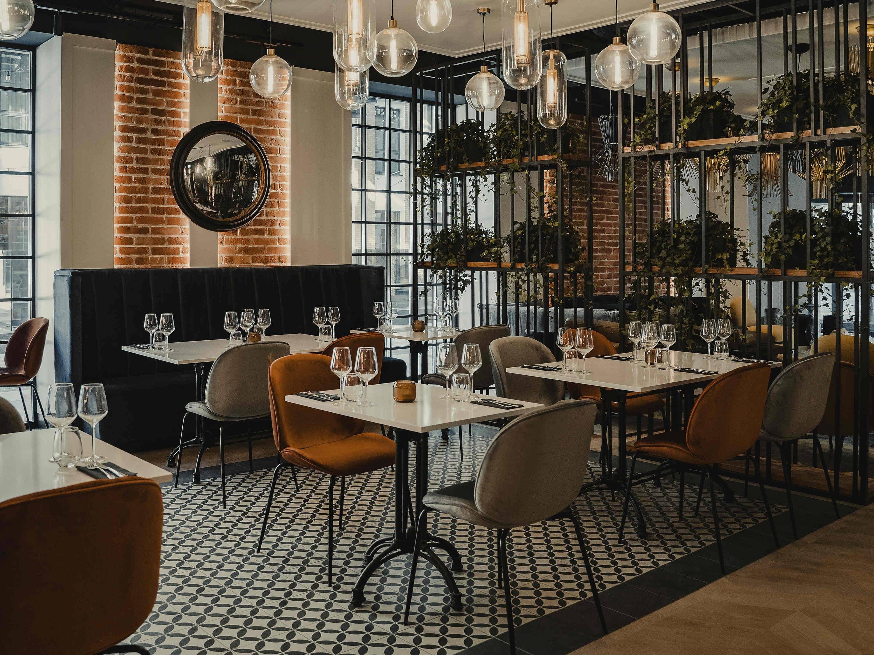 Interior de restaurante con paredes de ladrillo, amplias ventanas y lámparas colgantes. Las mesas están dispuestas con copas de vino y cubiertos.