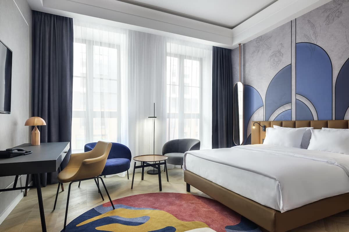Habitación de hotel con cama, escritorio, dos sillas, una mesita y un ventanal grande con cortinas.