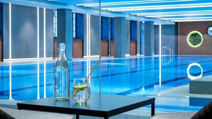Una piscina interior moderna con agua azul cristalina, una botella de vidrio con pajita y un vaso con un gajo de lima sobre una mesa.