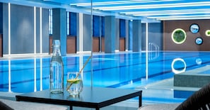 Una piscina interior moderna con agua azul cristalina, una botella de vidrio con pajita y un vaso con un gajo de lima sobre una mesa.