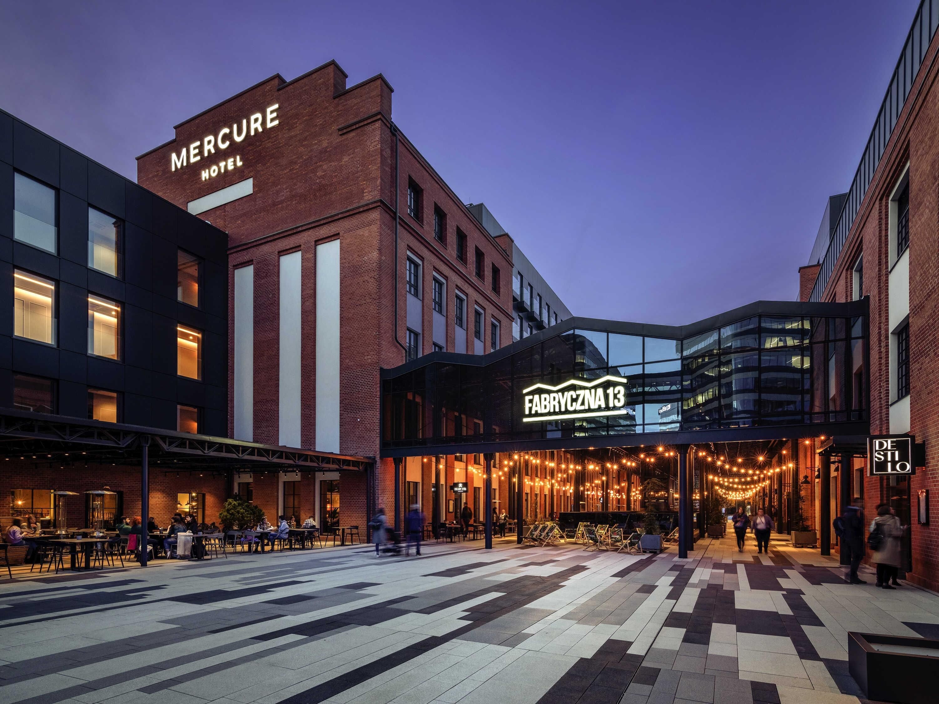 Hotellin yläpuolella on suuri, selkeästi näkyvä «Mercure Hotel» -kyltti, siinä on moderni lasinen sisäänkäynti ja ulkona on kivetty terassi, jossa on pöytiä ja tuoleja.