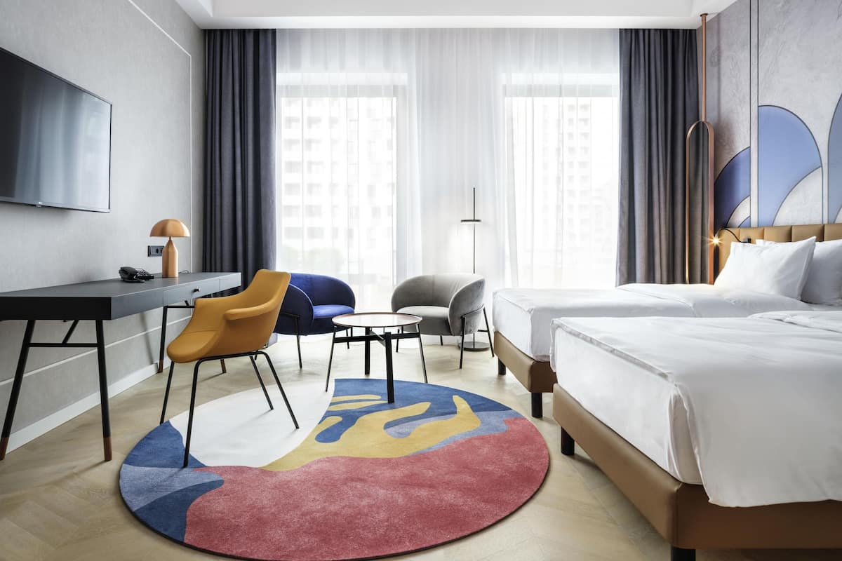 Habitación de hotel con cama, escritorio, silla, televisión y vista a edificios a través de amplias ventanas.