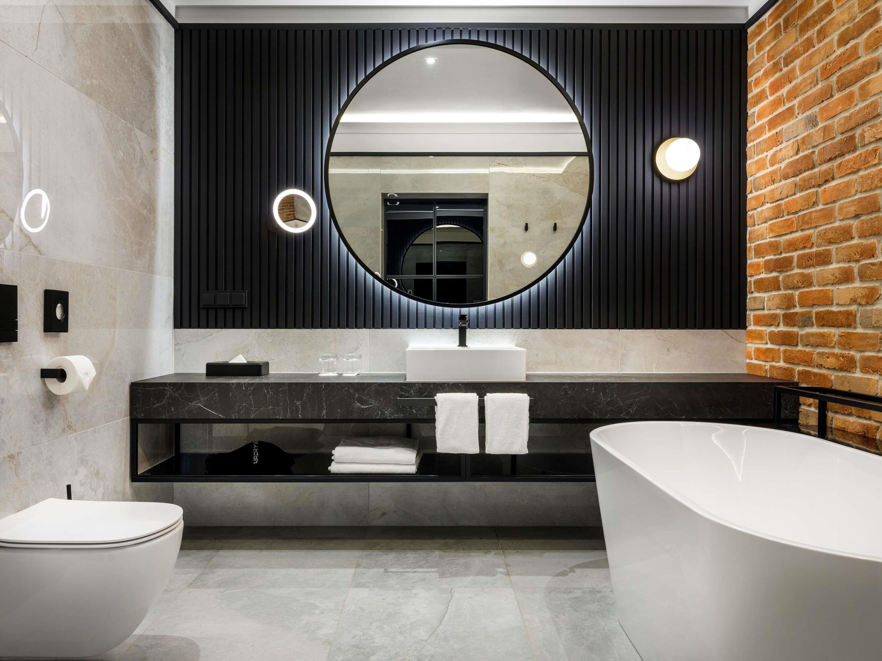 A modern bathroom with a round mirror, a black vanity, a white bathtub, and a toilet.