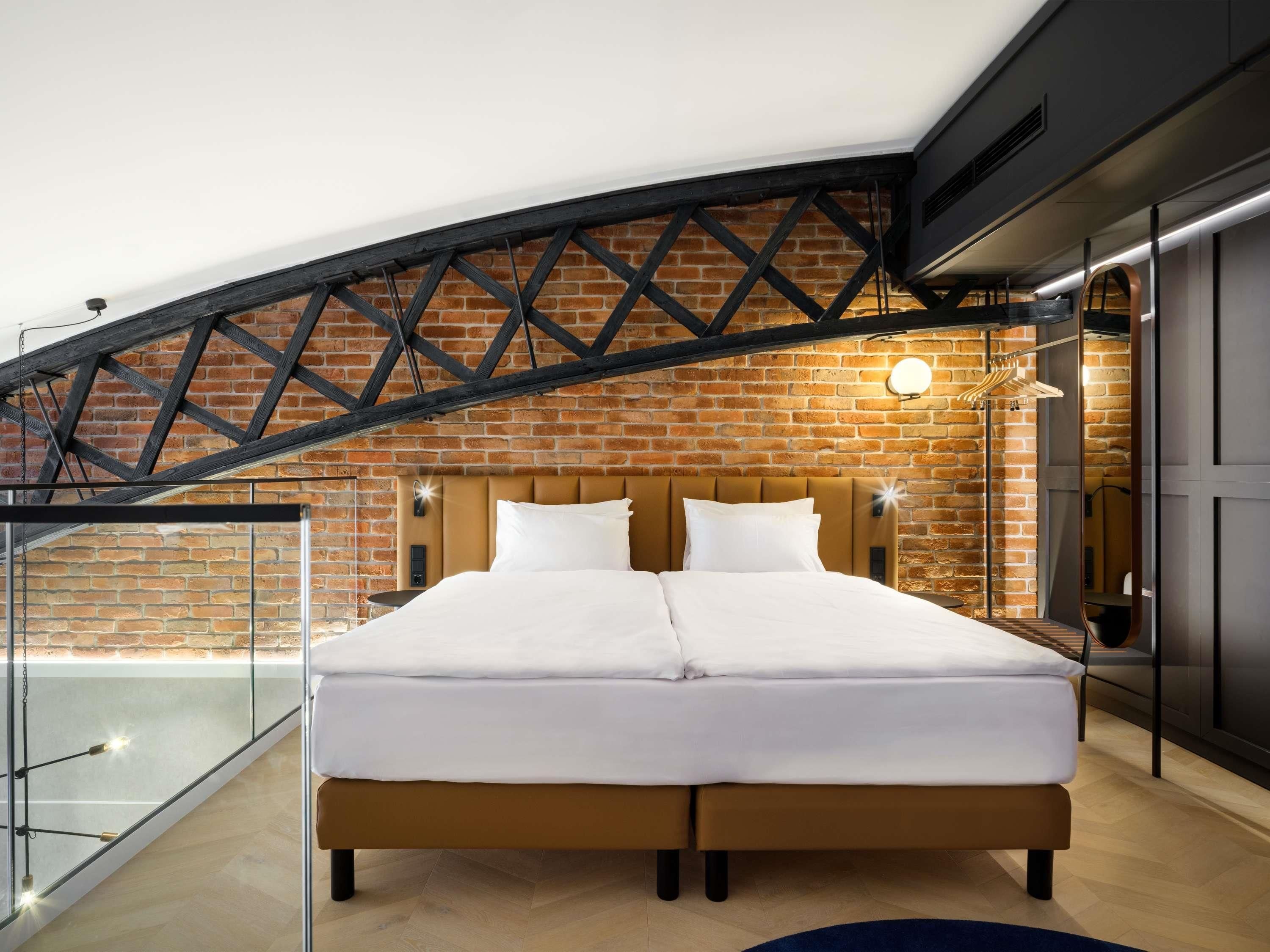 Un dormitorio con pared de ladrillo, cama con ropa blanca, cabecera de madera y balcón de cristal.