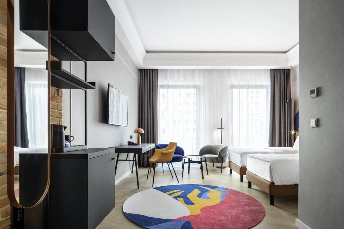 Una habitación de hotel moderna con cama, escritorio, silla y una alfombra colorida.