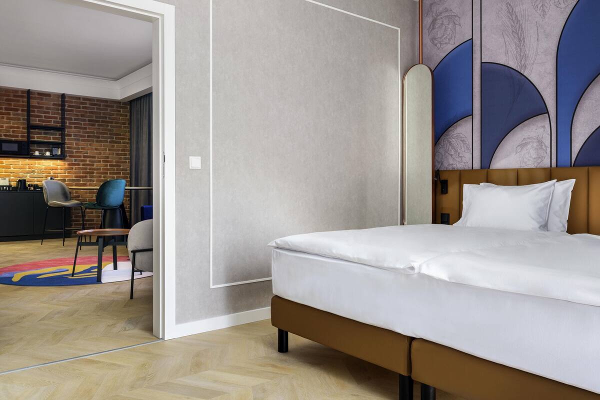 Habitación de hotel moderna con una cama grande, cabecera de madera y vistas a una zona de cocina con pared de ladrillo y bar.